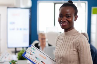 African woman manager looking at camera smiling, holding clipboard, while diverse coworkers talking in background. Manager working in professional start up financial business, modern company workplace ready for meeting.