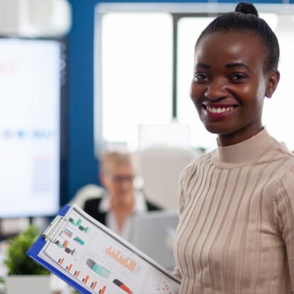 African woman manager looking at camera smiling, holding clipboard, while diverse coworkers talking in background. Manager working in professional start up financial business, modern company workplace ready for meeting.